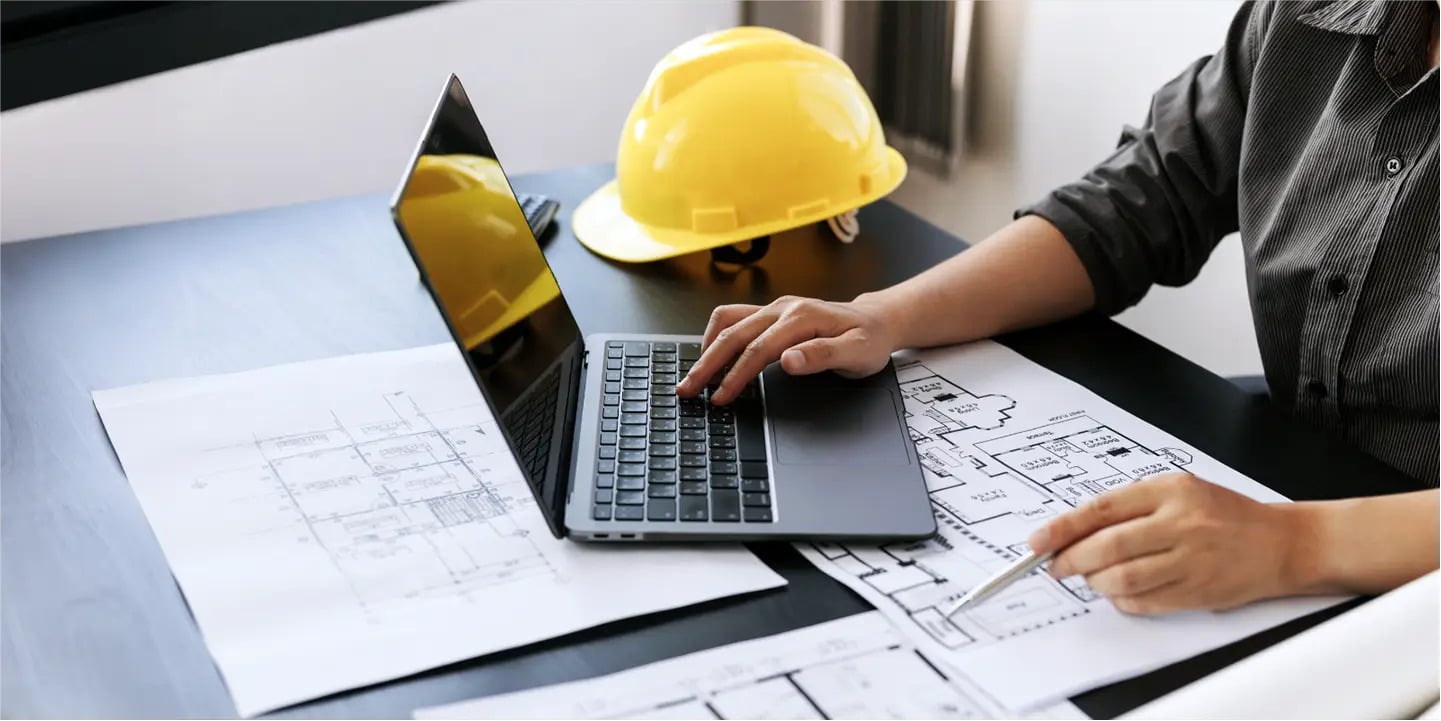 A person works on a laptop with architectural blueprints and a yellow hard hat on the desk.