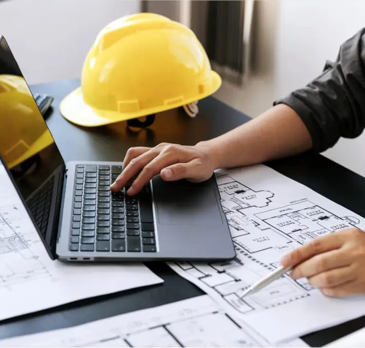 Person working on a laptop with architectural blueprints and a yellow construction helmet on the desk.