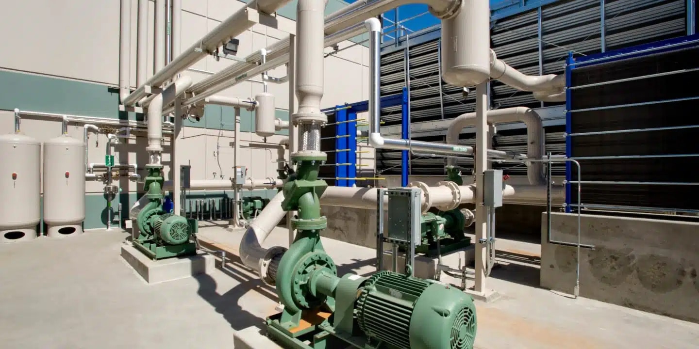 Industrial facility showing large pumps, pipes, and tanks in an outdoor mechanical area, likely part of a heating, cooling, or water management system.