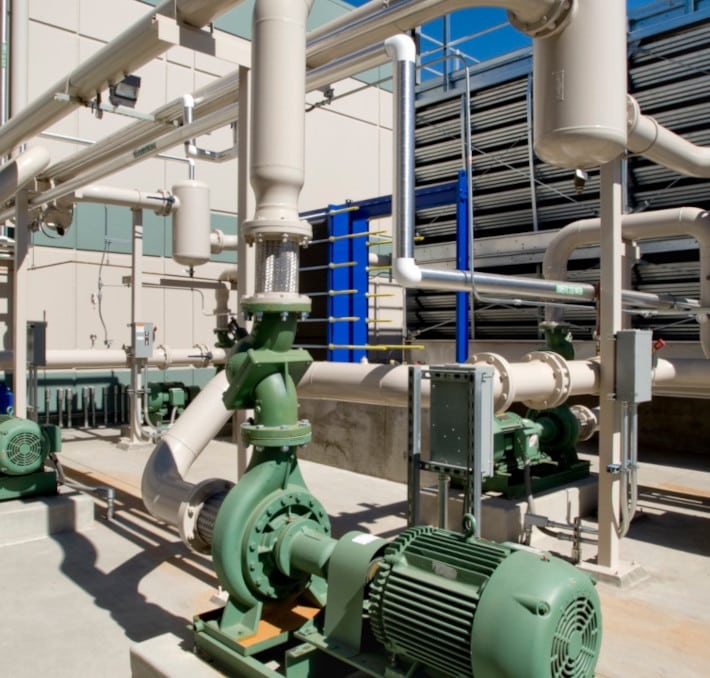 Industrial piping system with pumps, motors, and interconnected pipes outdoors, adjacent to a building and cooling units.