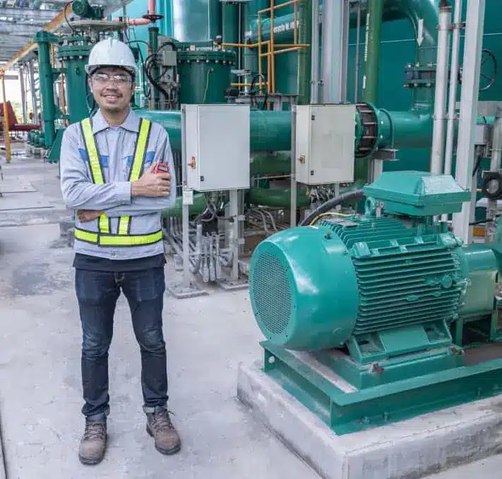 A worker wearing safety gear and a hard hat stands beside industrial machinery and large green pipes at a facility.