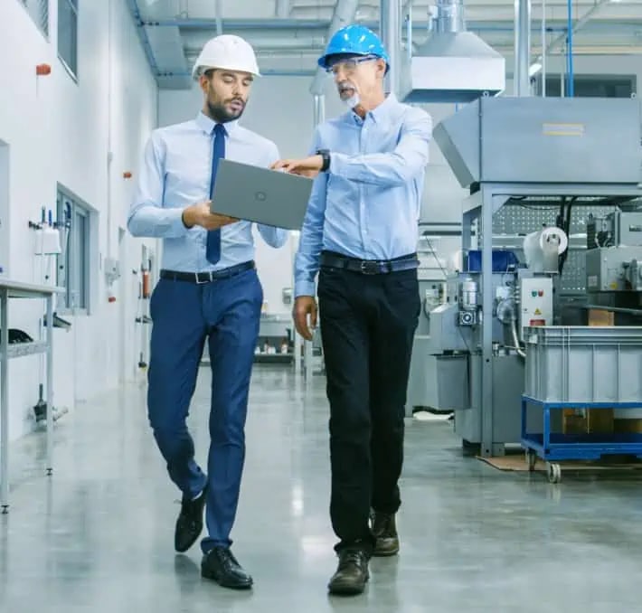 Two men wearing hard hats and business attire walk through a factory while looking at a laptop and discussing something. Industrial equipment is visible in the background.