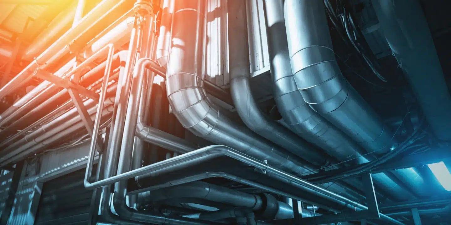 A network of large metal industrial pipes and ducts running along a ceiling, illuminated by bright lights.