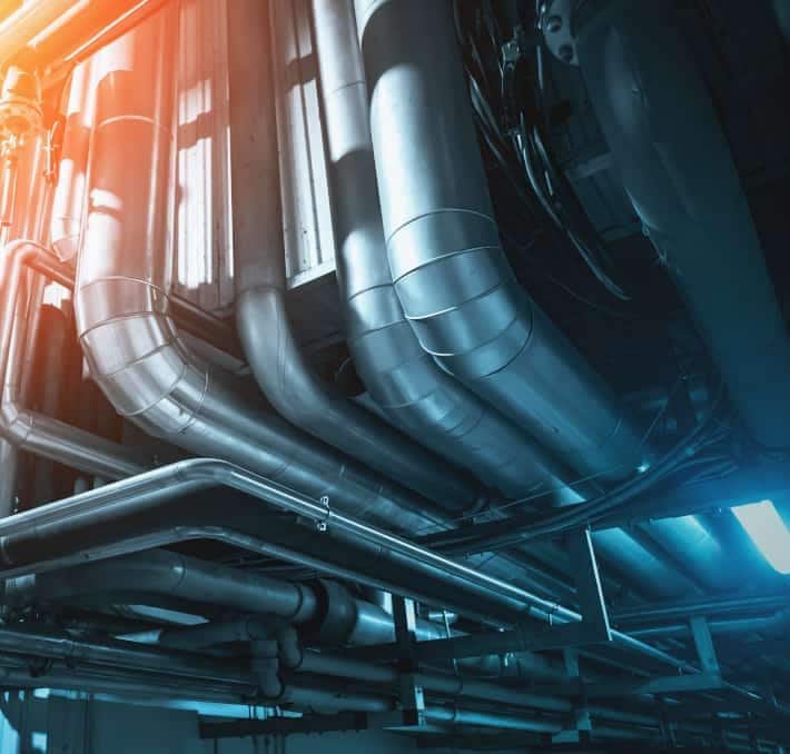 A network of large metal pipes and ducts runs across a ceiling in an industrial or mechanical facility, illuminated by blue and orange lighting.