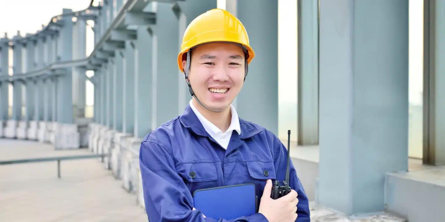 Man in blue work uniform and yellow hard hat smiles while holding a clipboard and walkie-talkie, standing outdoors near an industrial structure.