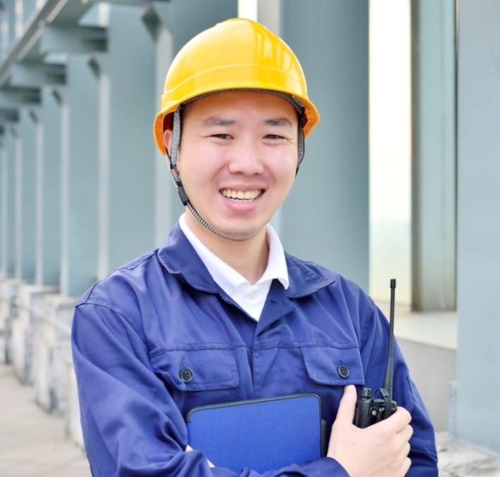 A person wearing a yellow hard hat and blue work uniform is smiling, holding a clipboard and a walkie-talkie, standing outdoors near a building.