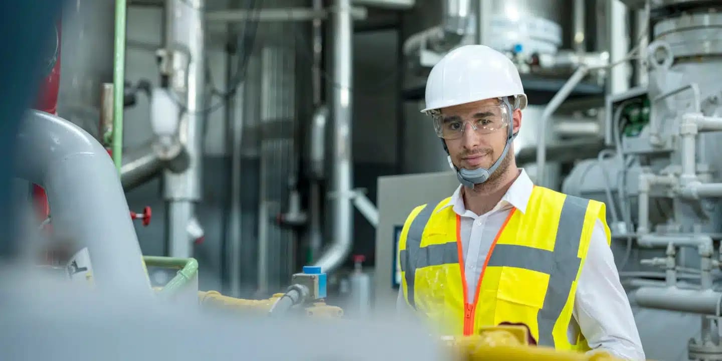 A man wearing a white hard hat, safety glasses, and a yellow reflective vest stands in an industrial facility with pipes and machinery in the background.