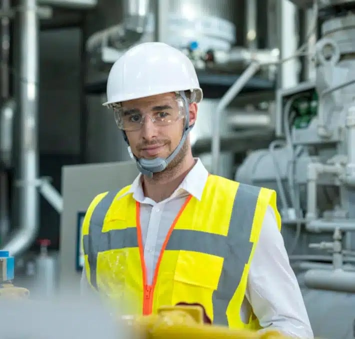 A man wearing a white hard hat, safety goggles, and a yellow reflective vest stands in an industrial setting with machinery in the background.