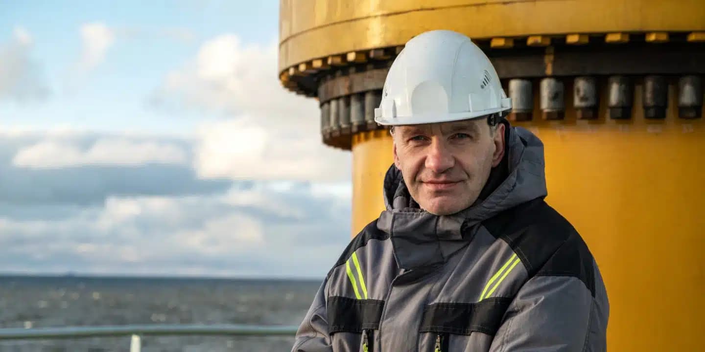 Man in a hard hat and protective jacket stands in front of a large yellow industrial structure by the sea, with cloudy sky in the background.