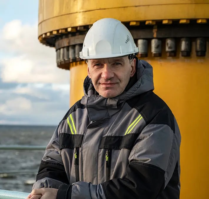 A man wearing a white hard hat and gray safety jacket stands outdoors near a large yellow industrial structure by the water.