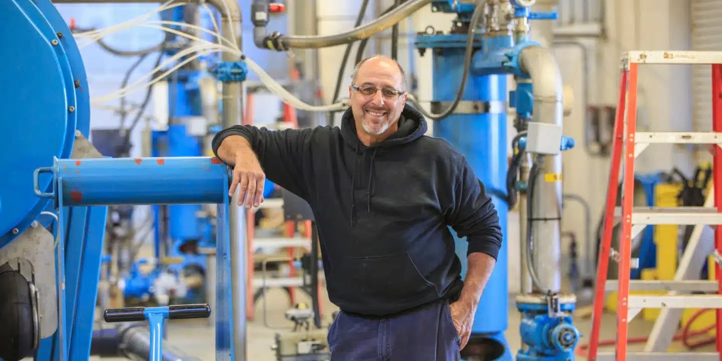 A man wearing glasses and a black hoodie stands and smiles in an industrial workshop with blue machinery and equipment in the background.