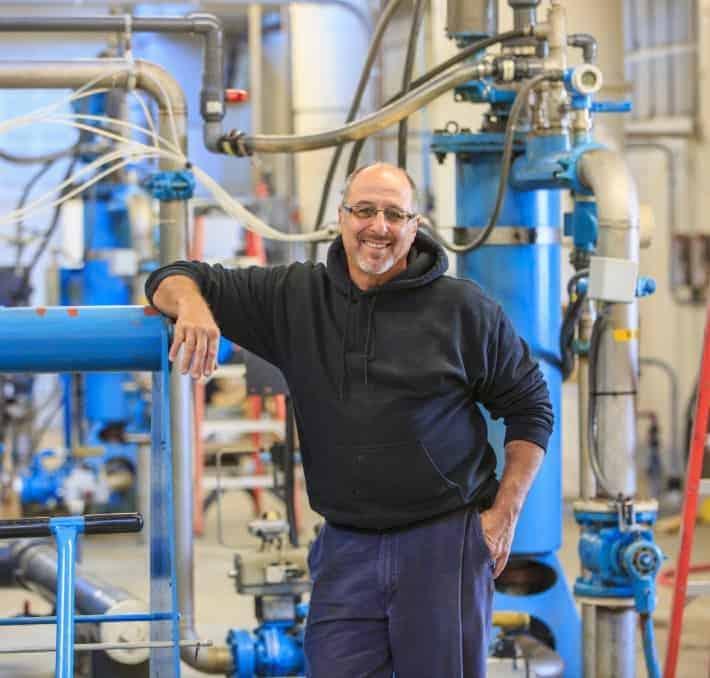 A man in a black hoodie stands smiling in an industrial facility with blue pipes and machinery in the background.