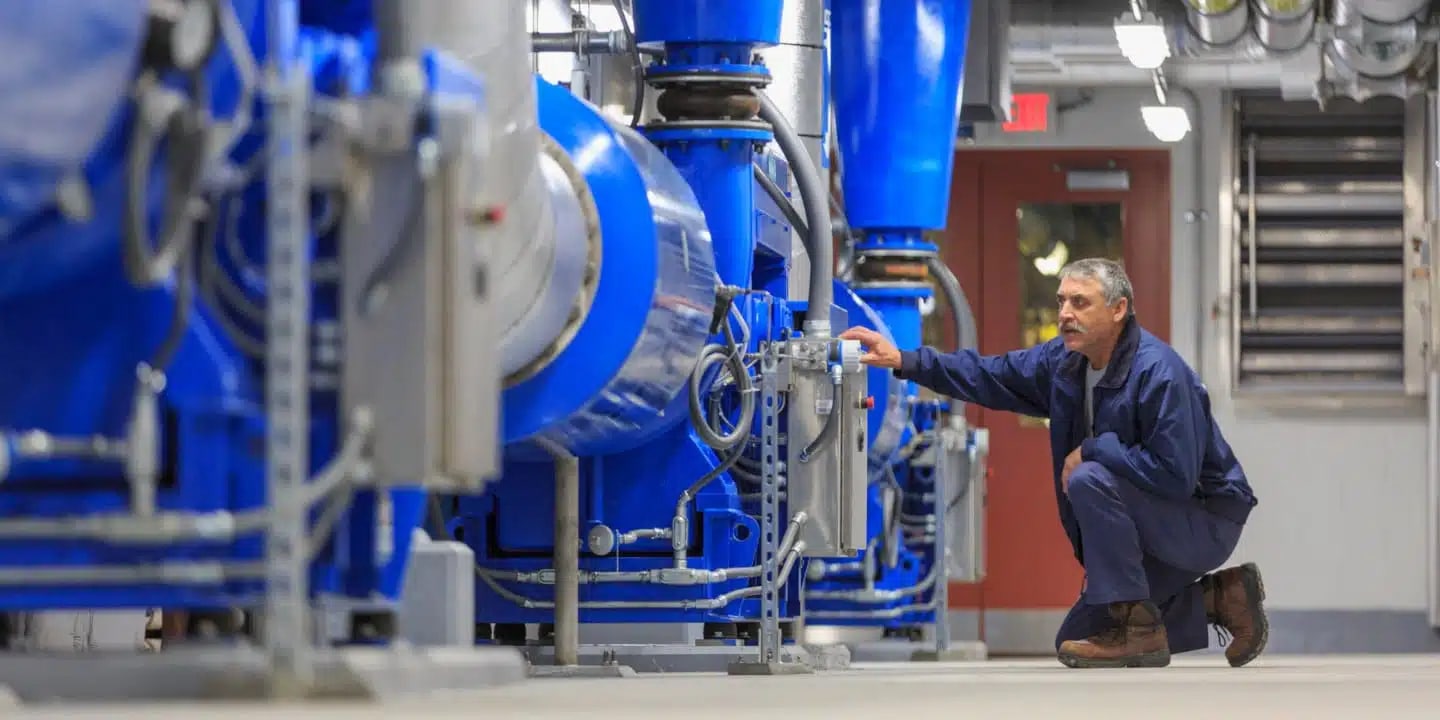 A man in work clothes inspects or adjusts a large blue industrial machine inside a clean, well-lit facility.