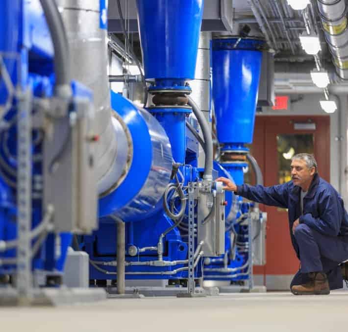 A man in work attire inspects large industrial blue machinery in a brightly lit facility.