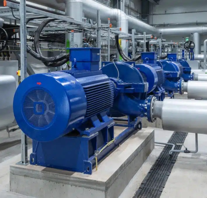 Blue industrial pumps and electric motors installed in a row inside a facility, surrounded by metal pipes and industrial equipment.