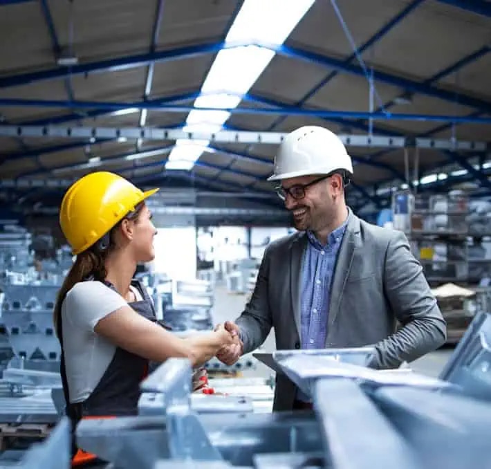Two people wearing hard hats shake hands inside a large industrial warehouse, surrounded by metal materials and equipment.