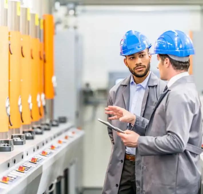 Two men in blue hard hats and lab coats discuss while inspecting industrial equipment with orange panels in a factory setting. One holds a tablet.